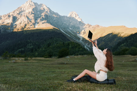 A joyful woman sits on a grassy meadow beneath rugged mountain peaks, lifting a laptop to capture a playful selfie against a clear blue sky.の写真素材