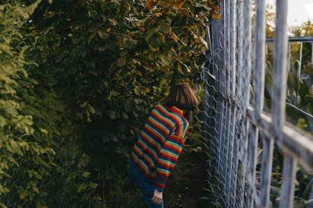 Child in a striped sweater peeking through a metal fence beside overgrown bushes, outdoor curiosity and exploration, young girl discovering a hidden garden path in natureの写真素材