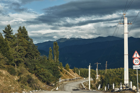 mountain road landscape with powerline and forest under cloudy sky on a rural asphalt curve, poles...