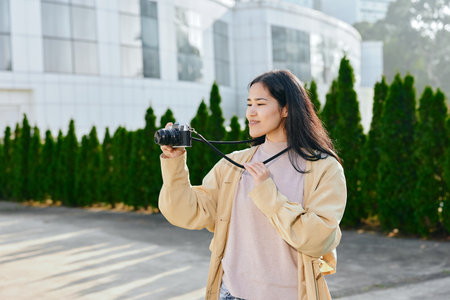 Woman holding a camera outdoors, casual jacket, soft sunlight, focused on framing a shot, greenery and modern architecture in the background, candid photography momentの写真素材