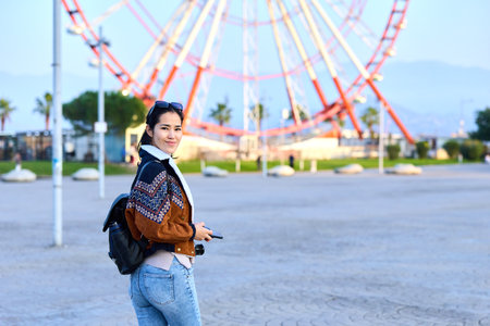 Woman with camera in outdoor amusement park beside a large ferris wheel, casual jacket, backpack, exploring and smiling as the scene captures travel and leisure momentsの写真素材