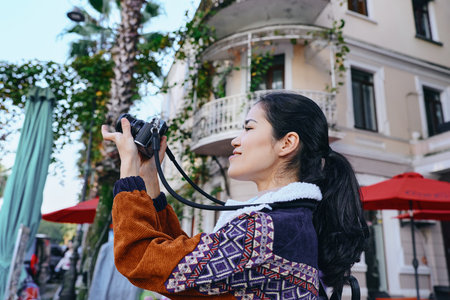 Photography of a woman photographer in a patterned sweater capturing urban street scenes with a camera, beside balconies and red umbrellas, showcasing travel, creativity, and street style.の写真素材