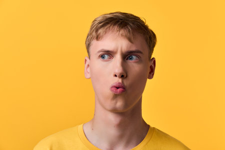 Man with a puzzled expression and pursed lips against a bright yellow background, showcasing curiosity and playful thinking in a casual sweater for lifestyle or reaction imageryの写真素材