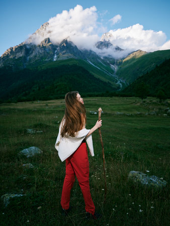 Woman in red pants holds a walking staff in a meadow, gazing toward towering mountains under a...