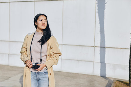 Woman with camera in casual fashion outdoors, standing confidently under bright sunlight, wearing beige coat and jeans, capturing candid street style and everyday moments.の写真素材