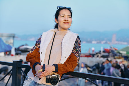 Woman leaning on a railing with a camera, wearing a plush brown faux fur jacket, by the water with a...