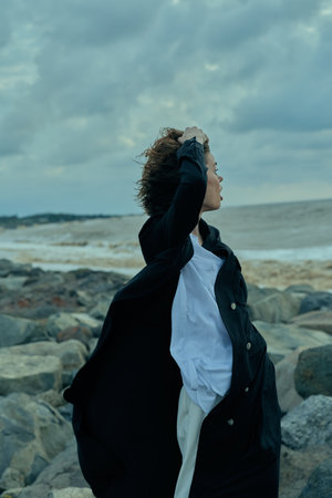 Woman on rocky shoreline wearing a dark coat and white shirt, hair blown by wind as she gazes toward...