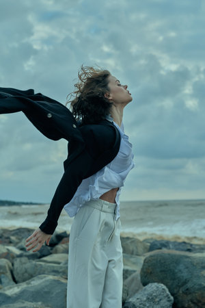 Woman on a rocky beach enjoys wind and motion, clothing billowing as sea and sky form a vast...