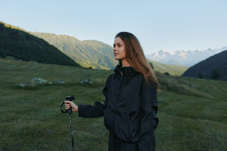 A lone female hiker stands in a green alpine meadow with a trekking pole. She wears a dark jacket...