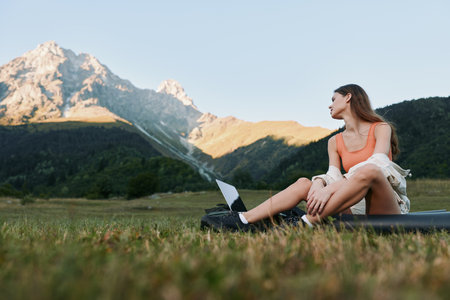 An outdoor scene shows a woman seated on grass with a laptop, gazing into the distance. She looks...