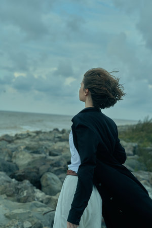 Woman standing on rocky coastline, wind blowing her hair, back turned, wearing a dress and dark...
