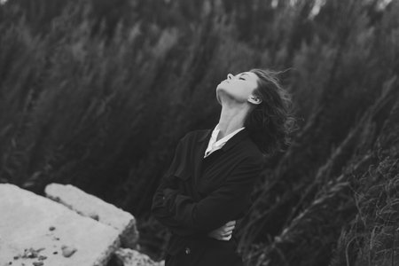 Contemplative woman gazing upward in a monochrome outdoor landscape, arms crossed, wind blowing through hair, solitary moment by rugged rocks, a moody study of resilience and natureの写真素材