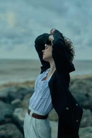 Windy coast fashion pose, woman lifts hair, wearing a white shirt and dark jacket, belt, standing...