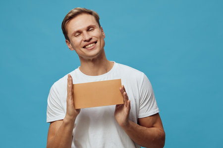 Smiling happy young man holding cardboard box with closed eyes isolated on blue background studio...