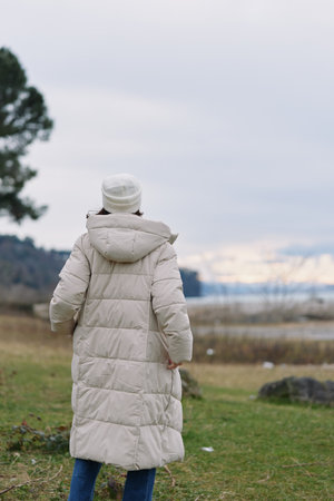 Person wearing a long beige winter coat stands in an open field with grass under a pale sky. The...