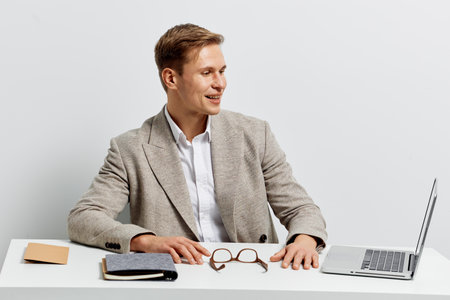 Smiling man in beige blazer sitting at desk with laptop and glasses in bright modern office environment, looking away from the camera. Business casual, professional appearance, concept of work, communication, success, positive attitude, modern lifestyle.の写真素材