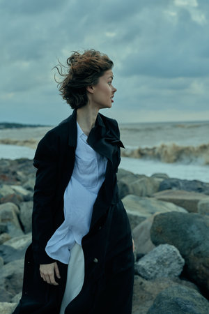 Windy coastal scene with a woman in a long coat standing among rocks as waves crash, creating a...