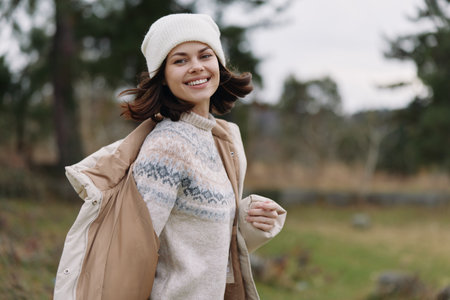 Woman in a cozy sweater and coat outdoors, smiling with a beanie, carrying a light jacket, autumn...