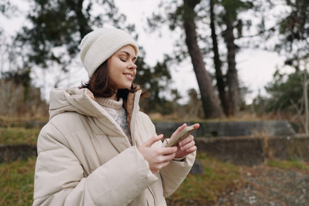 Woman in a warm beige coat and knit hat using a smartphone outdoors among trees, capturing a candid...