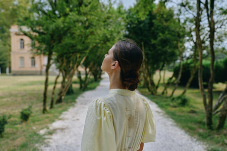 Woman seen from behind walking along a gravel path in a park, surrounded by greenery and trees,...