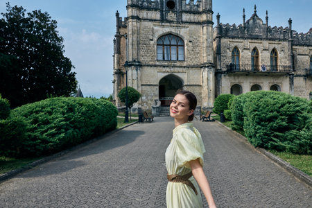 Woman in pale dress stands on a cobblestone path in front of a historic castle, smiling gently as lush greenery frames the grand architecture, and the scene exudes timeless charm.の写真素材