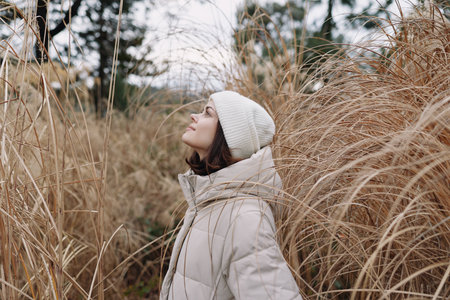 Woman in a light puffer coat and beanie gazes upward among tall grasses, capturing calm outdoor...