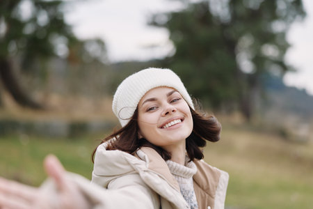 Portrait of a woman outdoors, smiling, reaching, in a beanie and casual jacket, captured in a sunny...