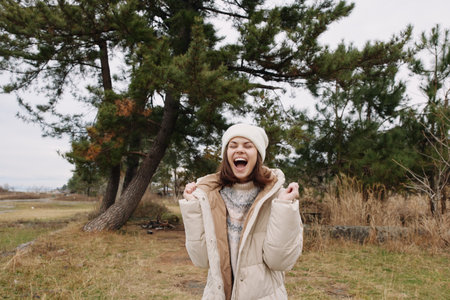 Outdoor woman in a padded coat and beanie celebrates with a bright smile in a windy field, trees in...