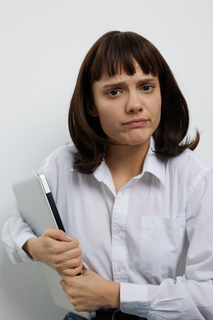 Portrait of a thoughtful young woman holding a notebook, looking serious. She wears a white shirt...