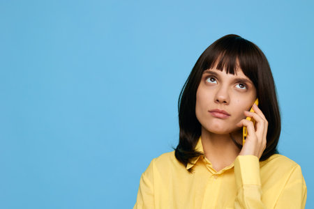 A thoughtful young woman wearing a yellow blouse speaks on a mobile phone against a bright blue...