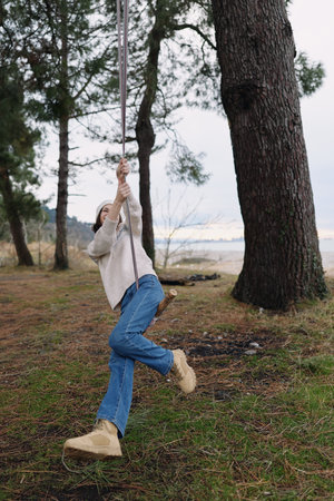 Person on a rope swing in a forested park beside a lake, casual outfit with jeans and boots, capturing a playful outdoor momentの写真素材