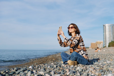 Woman taking a selfie on a rocky beach, wearing a plaid shirt and jeans, sunglasses on, close to the...