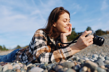 Woman photographer lying on pebbles outdoors, smiling and focused, holding a camera in a casual plaid shirt, enjoying nature and a creative photography moment by the shoreの写真素材