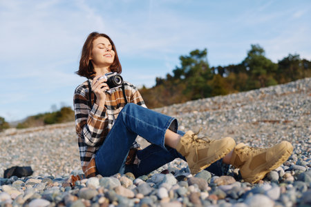 Woman photographer on a rocky beach smiles with a camera, relaxed pose under blue sky, capturing outdoor scenery and nature during a casual photo session.の写真素材