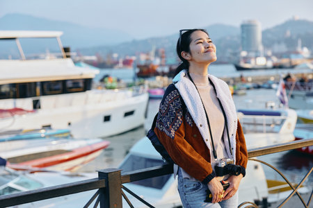 Smiling woman wearing a thick knit jacket on a harbor pier with boats and water in the background,...