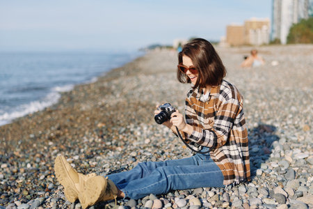Woman on a pebbled beach with a camera, wearing plaid shirt and denim, enjoying sunny outdoor...