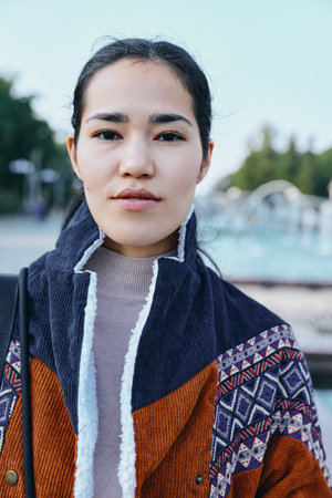 Woman outdoors wearing a patterned coat over a light knit sweater near a pool, calm expression,...