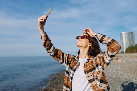 Woman by the seaside taking a selfie with a smartphone, outdoors on a sunny day, wearing a plaid...