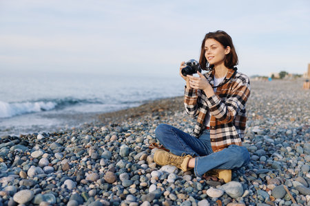 Woman photographer sits on a pebble beach, holding a camera and capturing seaside scenery in a...