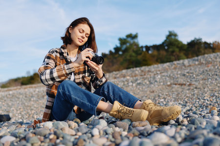 Woman sitting on a pebble beach, outdoors, holding a camera for photography, wearing a plaid shirt and jeans with boots, focused on a shot under clear sky and natural lightの写真素材
