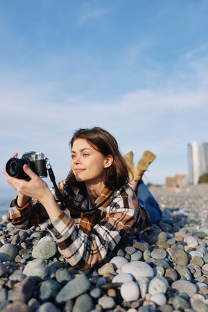 Female photographer lying on a rocky beach, examining a camera, plaid shirt, sunny day, distant...