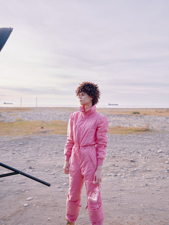 Pink jumpsuit worn by a model in an outdoor coastal setting, portrait style with wind in hair, soft...