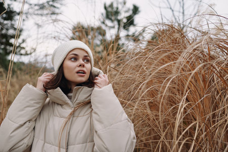 Woman in a beige puffer coat and knit beanie adjusting her hood in a chilly outdoor setting among...