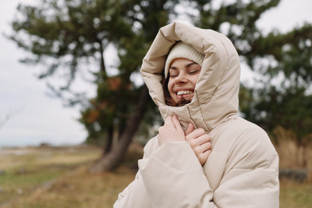 Woman in a puffy hooded coat outdoors, smiling warmly amid trees and an open field, in chilly...