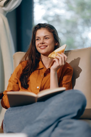 Happy young woman enjoying a book and a slice of pie in a cozy home setting, wearing an orange shirt...