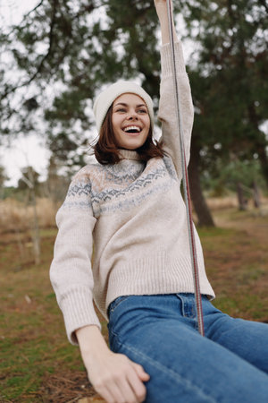 Joyful woman on a swing outdoors in a cozy knit sweater and beanie, laughing as she holds the ropes in a park setting with trees and soft natural light.の写真素材