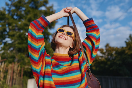 Woman in a colorful striped sweater and yellow sunglasses smiling outdoors; young adult joyful and carefree, raising a shoulder bag strap in a park scene for casual fashion travel lifestyle.の写真素材