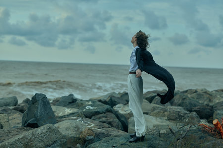 Figure stands on rocky shoreline with cape flowing in wind, eyes toward the horizon, creating a...