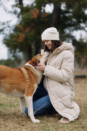 Woman kneels in a beige coat and beanie, petting a dog in an outdoor setting, sharing a calm,...