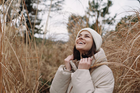 Woman in winter coat and beanie enjoying outdoor nature among tall grasses, smiling up with a warm...